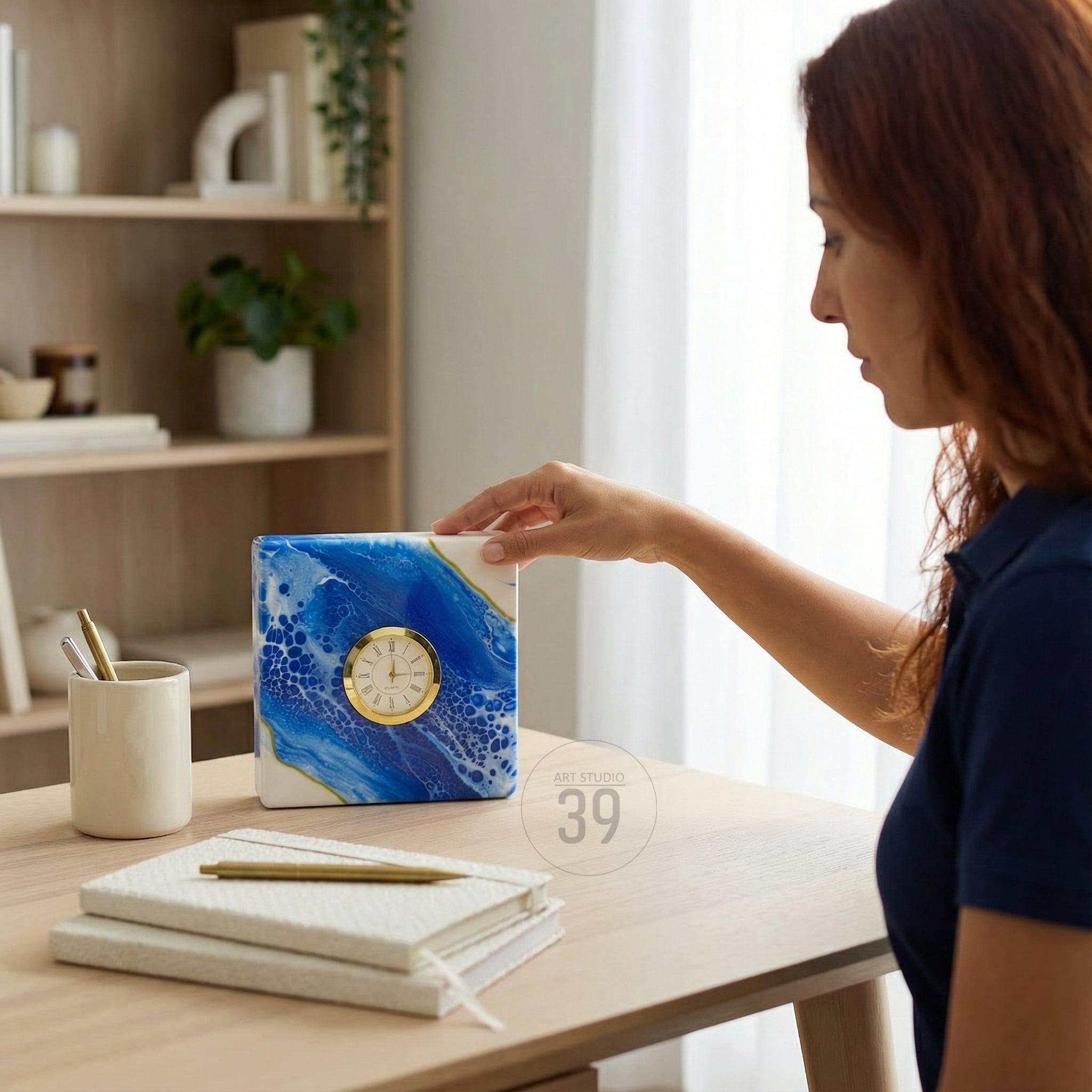 A square clock with a gold-colored clock face and hands, set against a blue and white abstract design. The clock is on a wooden surface, with a pen, notebooks, and a cup nearby. The clock has the text "ART STUDIO 39".