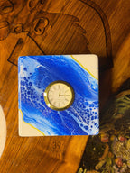 Square clock with a white base and a blue and white abstract design. The clock face is gold with Roman numerals. The clock is sitting on a wooden surface. The number "39" is visible on the bottom corner.