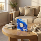 A square clock with a white and blue abstract design, featuring a gold-colored clock face. The clock is on a wooden coffee table, with a beige sofa and pillows in the background. The number "39" is visible on the bottom right.
