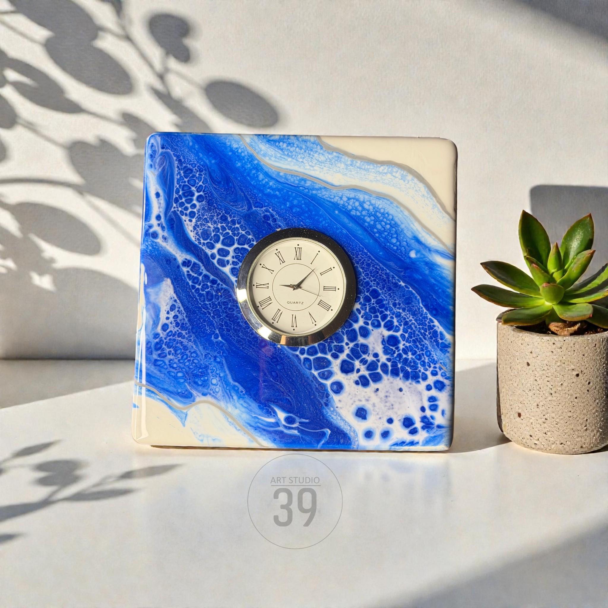 A square clock with a blue and white abstract design, featuring a silver-rimmed clock face with Roman numerals. The clock is next to a small potted succulent. The text "ART STUDIO 39" is at the bottom.