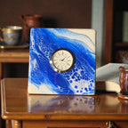 A square quartz clock with a white face and Roman numerals. The clock is set in a blue and white resin base with a wave-like pattern. The clock is approximately 4 inches wide.