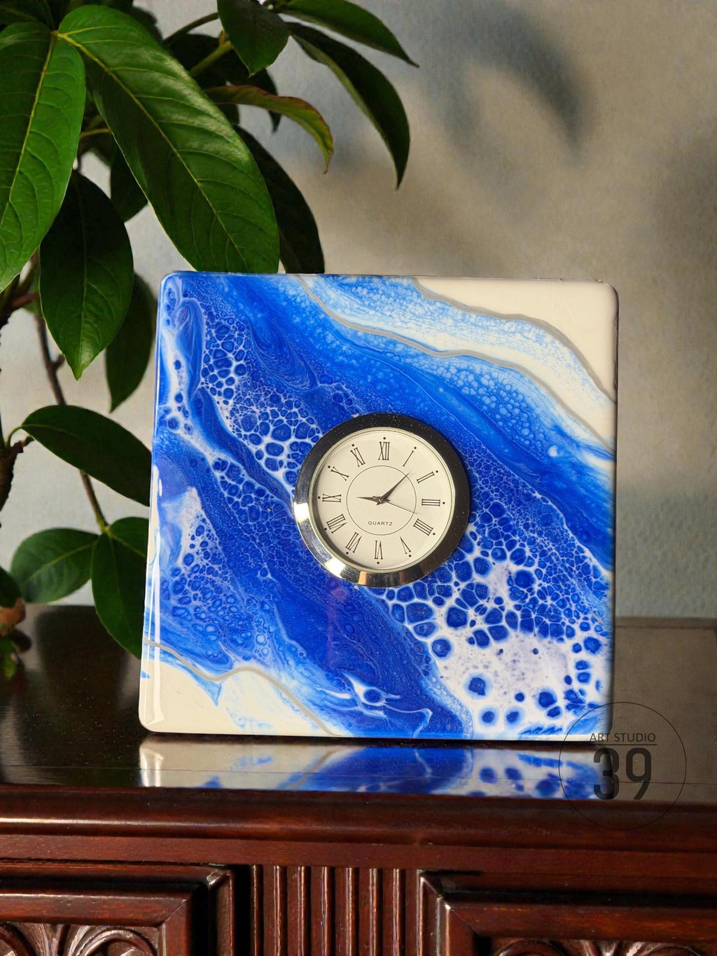 Square quartz clock with a blue and white abstract design. The clock face is white with black Roman numerals and hands. The clock is labeled "ART STUDIO 39".
