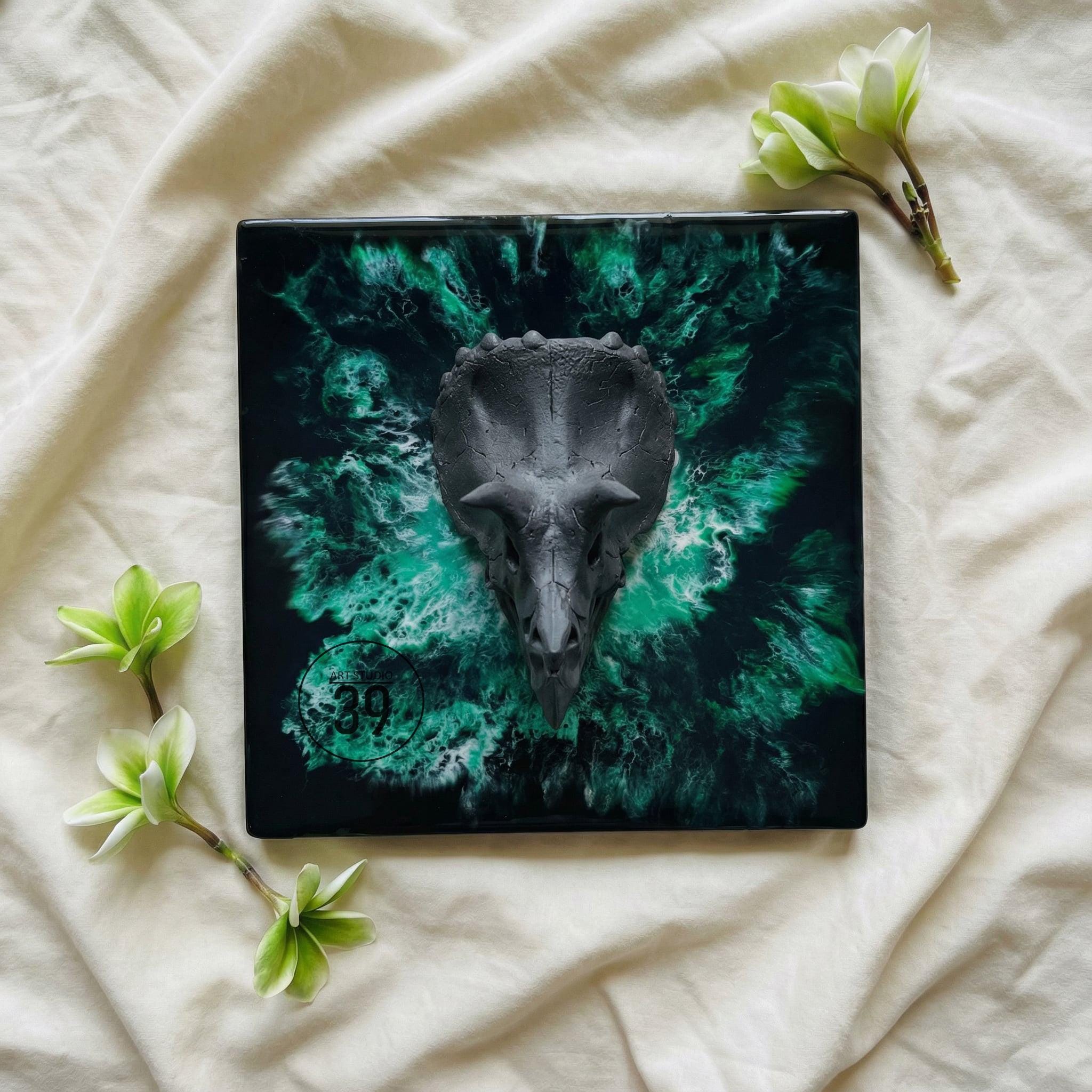 A square, black decorative piece featuring a gray triceratops skull. The background has a swirling green and black design. The number "39" is visible in the lower left corner. Green flowers are placed around the piece.