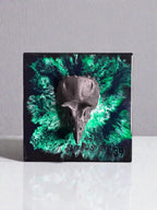 A square, black resin art piece featuring a dark gray bird skull emerging from a vibrant green and black abstract design. The artwork has a glossy finish and a small, circular label with the number "39".