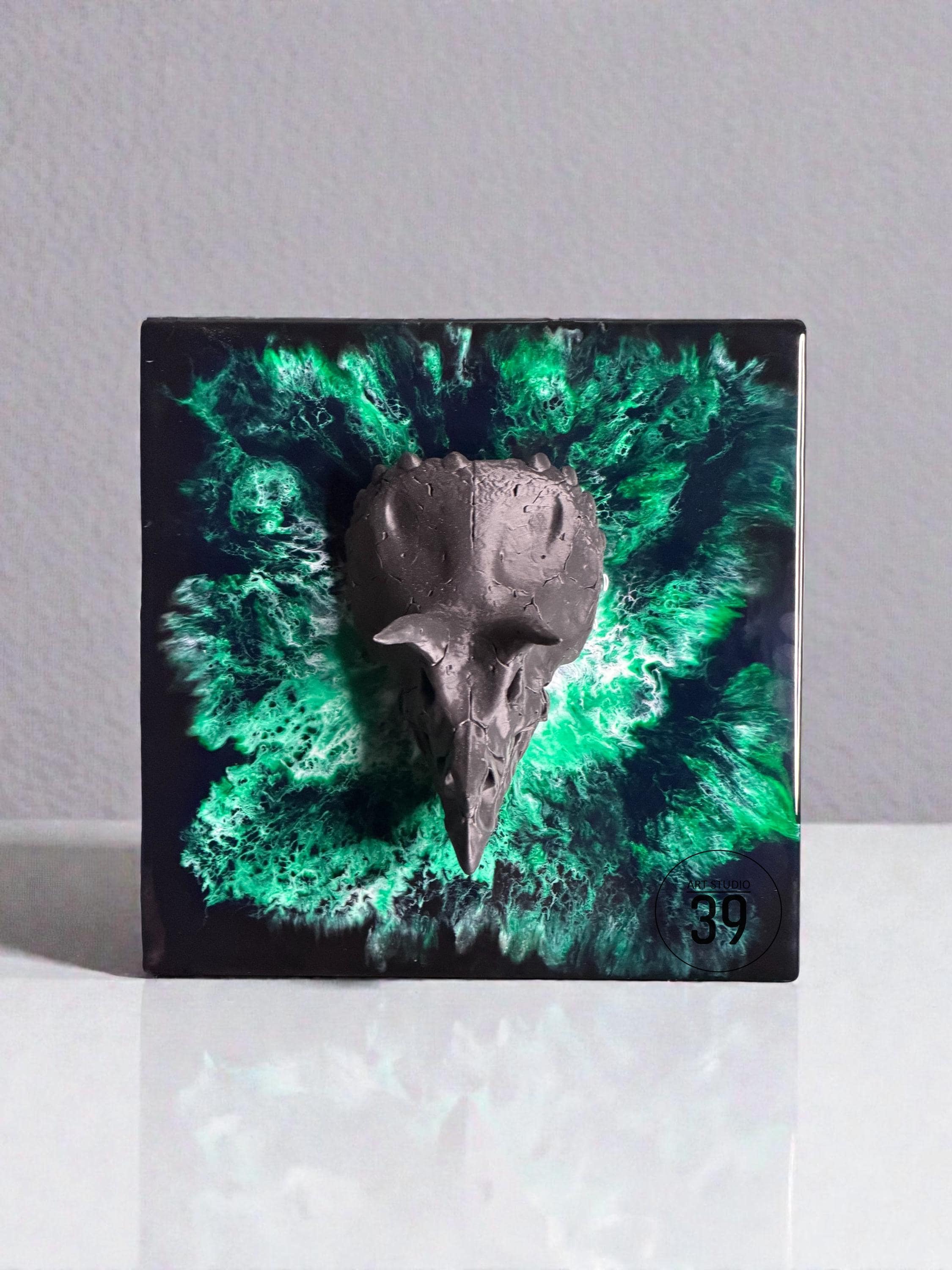 A square, black resin art piece featuring a dark gray bird skull emerging from a vibrant green and black abstract design. The artwork has a glossy finish and a small, circular label with the number "39".