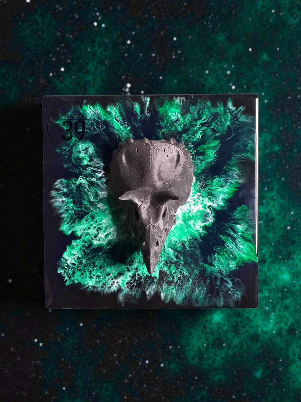 A dark gray sculpture of a bird skull is centered on a square, black base with a vibrant green and black abstract design. The number "39" is visible in the upper left corner. The background is a dark green, starry pattern.