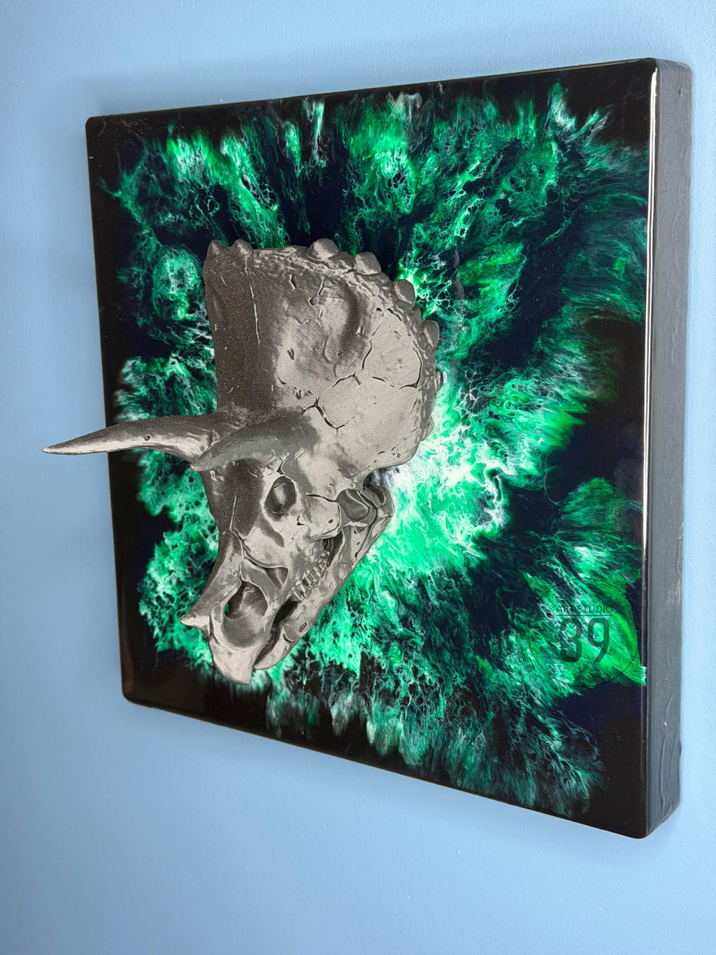 A metallic gray Triceratops skull sculpture mounted on a square black and green abstract background. The artwork features a textured skull with horns and a vibrant, swirling green and black design.