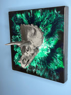 A metallic gray Triceratops skull sculpture mounted on a square black and green abstract background. The artwork features a textured skull with horns and a vibrant, swirling green and black design.