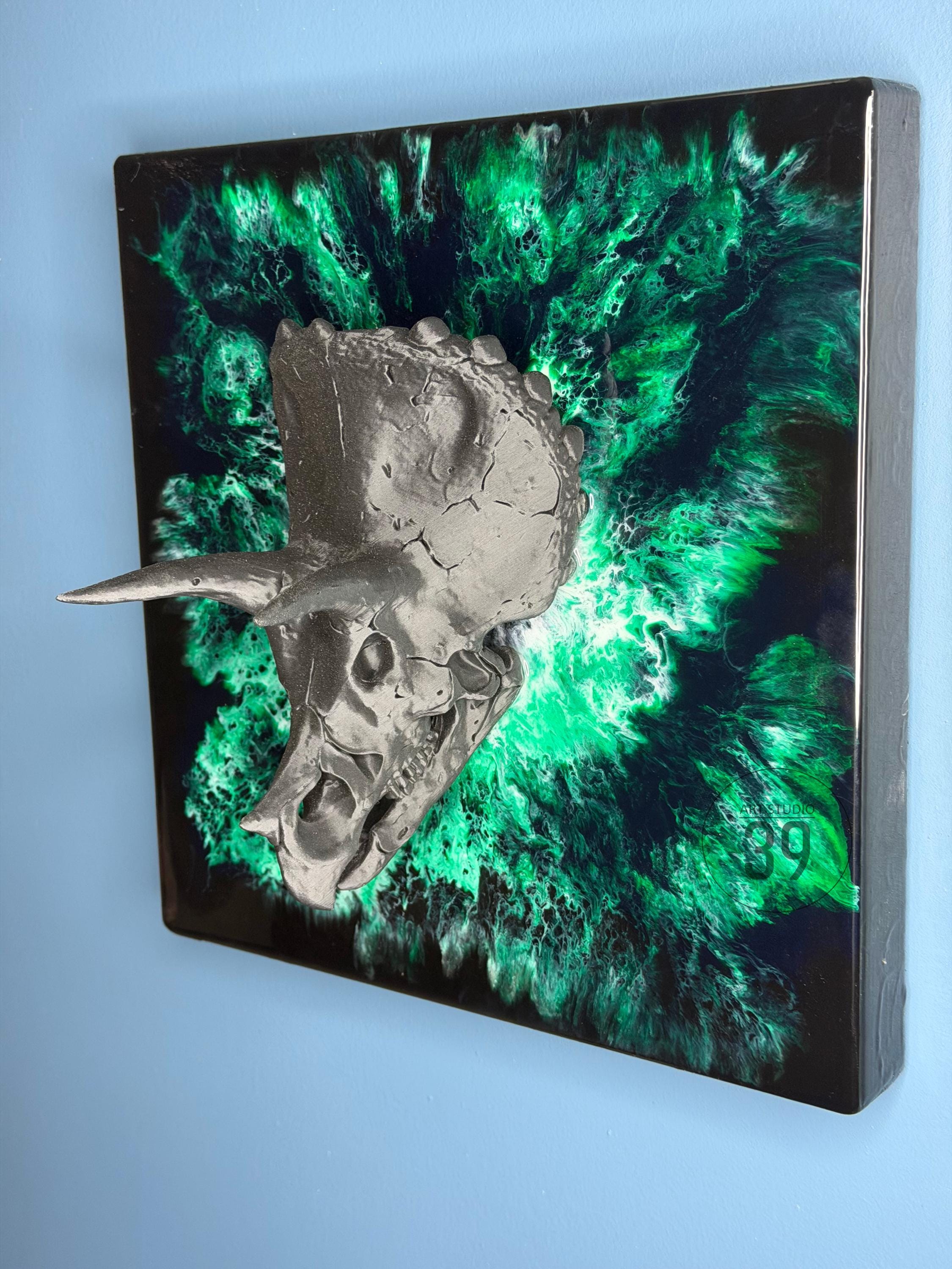 A metallic gray Triceratops skull sculpture mounted on a square black and green abstract background. The artwork features a textured skull with horns and a vibrant, swirling green and black design.