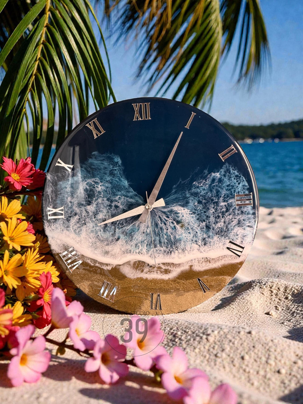 Round wall clock with a beach-themed design. The clock face features a dark blue ocean scene with white waves and a sandy beach. Roman numerals mark the hours. The clock is displayed on a sandy surface with flowers.