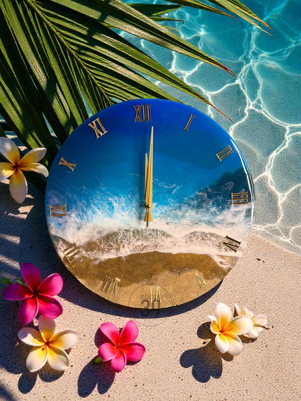 A round wall clock with a beach scene design. The clock face features a blue ocean, white waves, and a sandy beach. Gold Roman numerals mark the hours. The clock is surrounded by pink and white flowers.