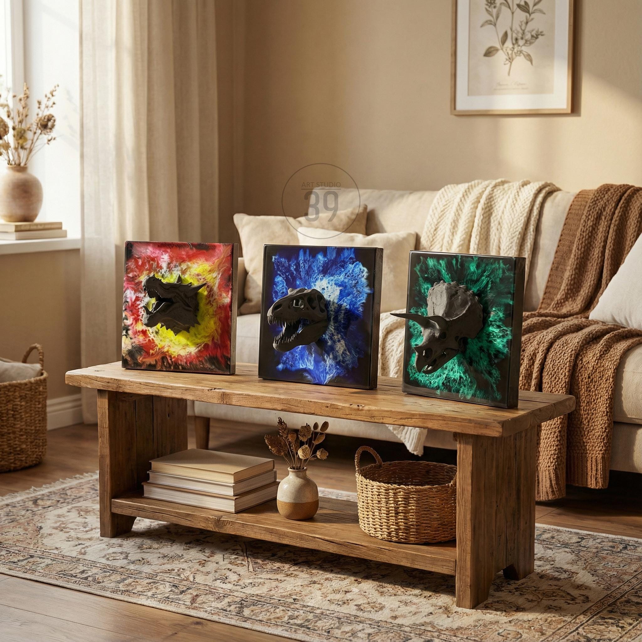 Three framed dinosaur skull art pieces. Each features a black dinosaur skull against a colorful background: red and yellow, blue, and green. The artwork is displayed on a rustic wooden coffee table.