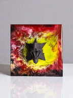 A square artwork featuring a dark gray, three-dimensional, stylized creature head. The background has a swirling pattern of red, yellow, and black. The artwork is on a white surface.