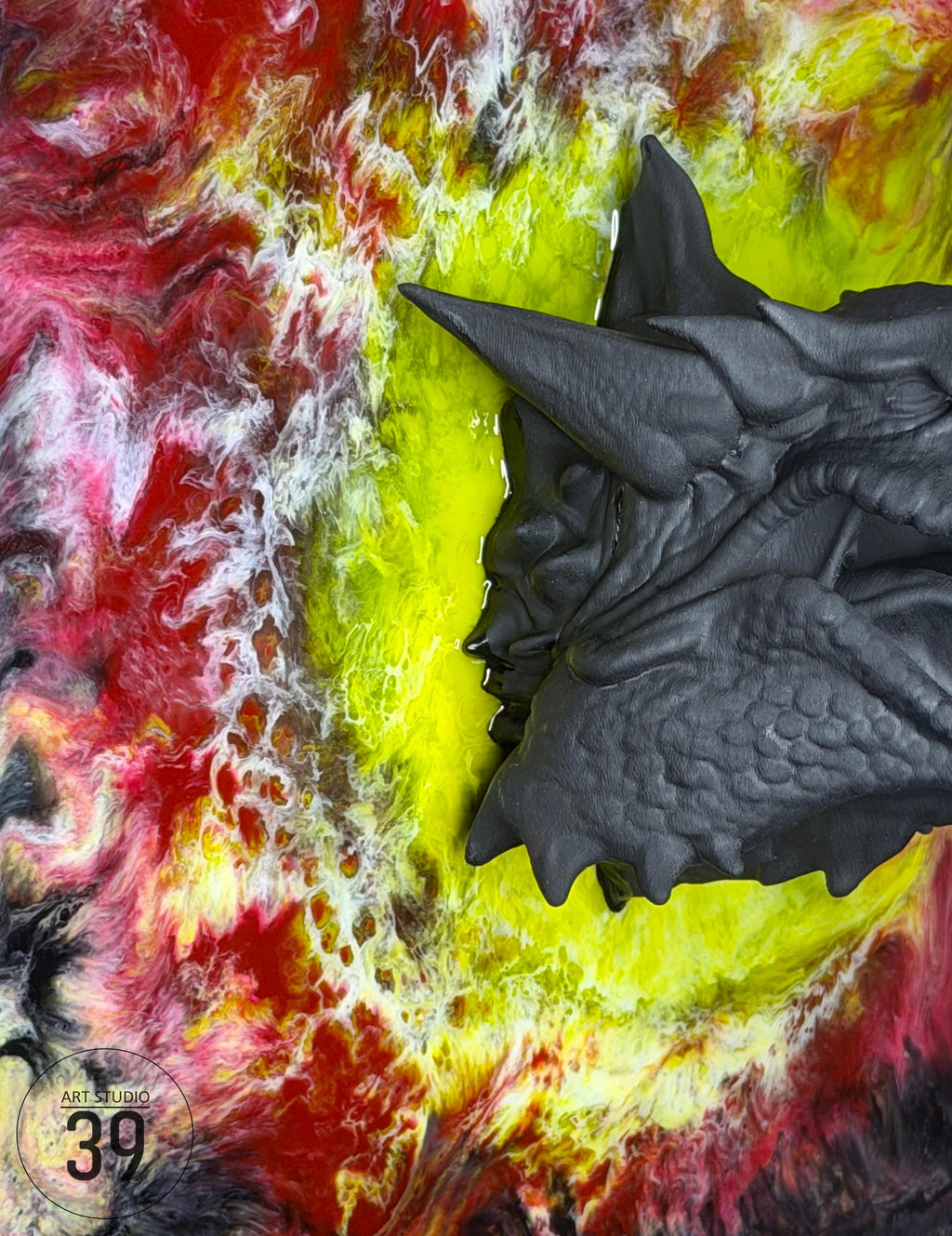 A dark gray, textured sculpture of a dragon's head with sharp horns and detailed scales. The dragon head is set against a vibrant background of swirling red, yellow, and white resin art. The artwork is signed "ART STUDIO 39".