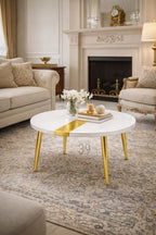 A white round coffee table with gold-colored legs and a gold stripe across the top. The table is in a living room setting with a rug, sofa, and fireplace.