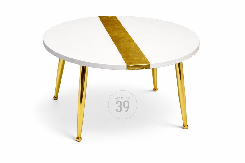 A round white coffee table with a gold stripe down the center. The table has four gold-colored legs. The table is a modern design and is suitable for a living room or lounge area.