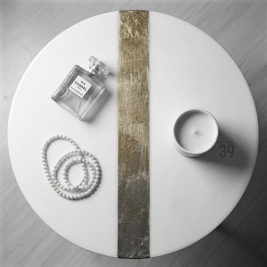 A white, round table with a gold and silver stripe down the center. A bottle of Chanel No. 5 perfume, a strand of pearls, and a small white candle are arranged on the table. A small white circle with the number 39 is also on the table.