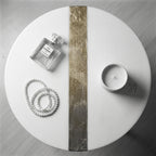 A white, round table with a gold and silver stripe down the center. A bottle of Chanel No. 5 perfume, a strand of pearls, and a small white candle are arranged on the table. A small white circle with the number 39 is also on the table.