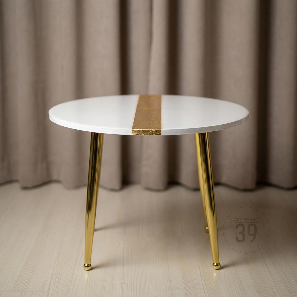 A round white side table with gold-colored legs and a gold stripe down the center of the tabletop. The table has a modern design and is suitable for a living room or bedroom. The table is approximately 20 inches in diameter.