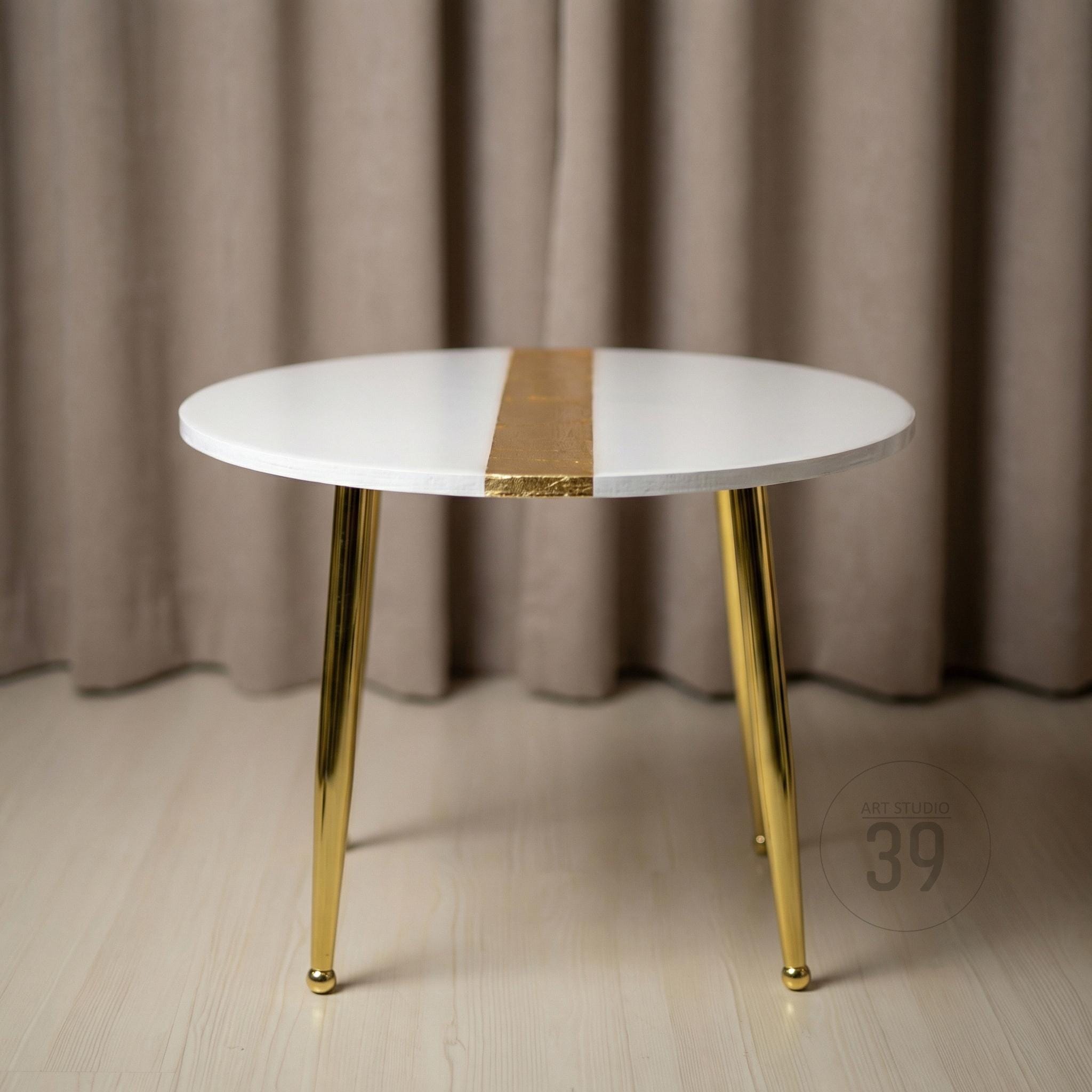 A round white side table with gold-colored legs and a gold stripe down the center of the tabletop. The table has a modern design and is suitable for a living room or bedroom. The table is approximately 20 inches in diameter.
