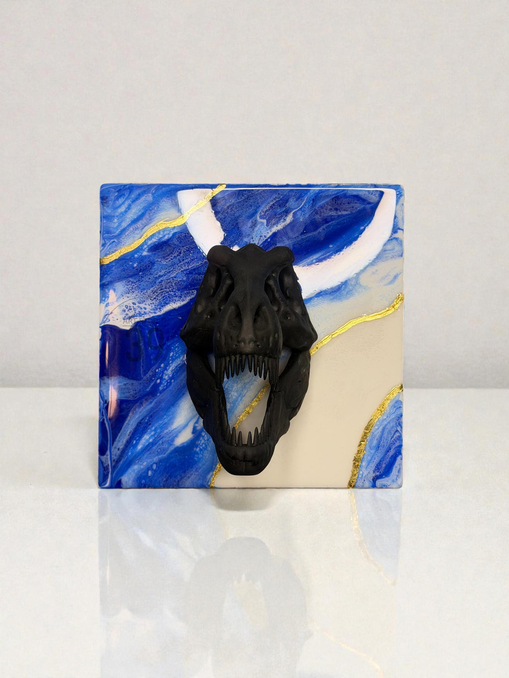 A square decorative piece featuring a black Tyrannosaurus Rex skull over a blue and white marbled background with gold accents. The piece has a glossy finish and a white border.