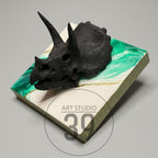 A black triceratops skull sculpture rests on a square, resin-coated surface. The surface features a white and green marbled design with gold accents. The sculpture is a unique piece of art.