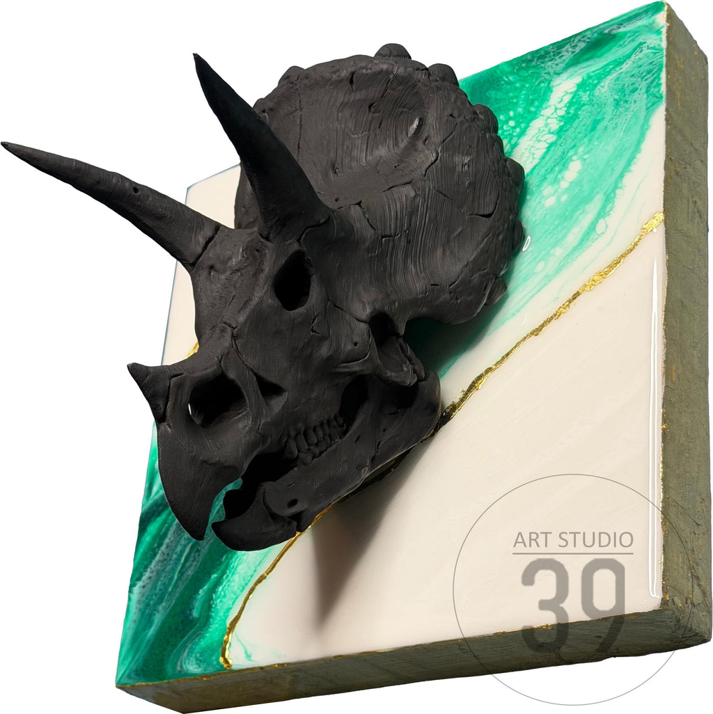 A black triceratops skull sculpture mounted on a square, abstract background. The background features a diagonal split with green and white sections, accented by a gold line. The art piece is labeled "ART STUDIO 39".