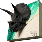 A black triceratops skull sculpture mounted on a square, abstract background. The background features a diagonal split with green and white sections, accented by a gold line. The art piece is labeled "ART STUDIO 39".