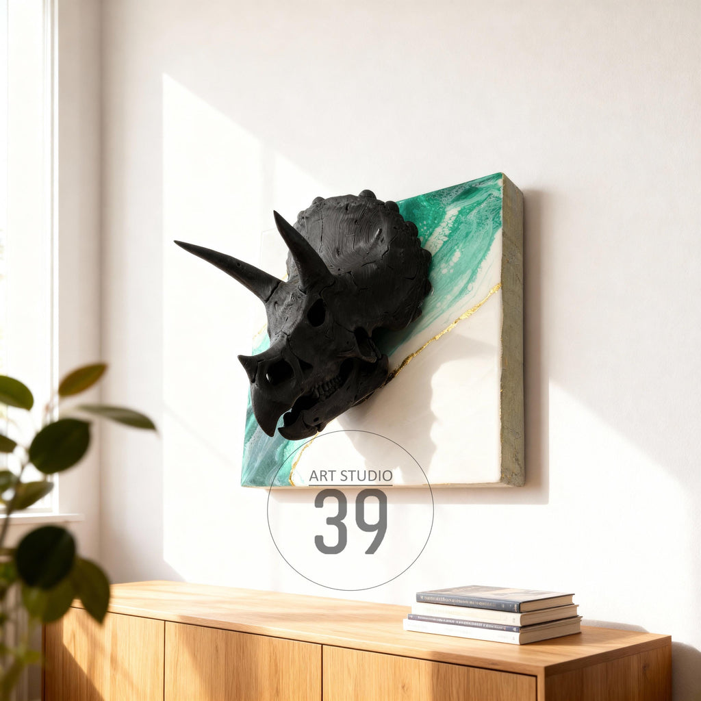 A black triceratops skull sculpture mounted on a square canvas. The canvas features a white background with green and gold accents. The artwork is displayed on a white wall above a wooden cabinet. The text "ART STUDIO 39" is visible.