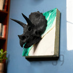 A black triceratops skull sculpture mounted on a square canvas. The canvas features a white and green abstract design with gold accents. The words "ART STUDIO 39" are printed on the lower right corner of the canvas.