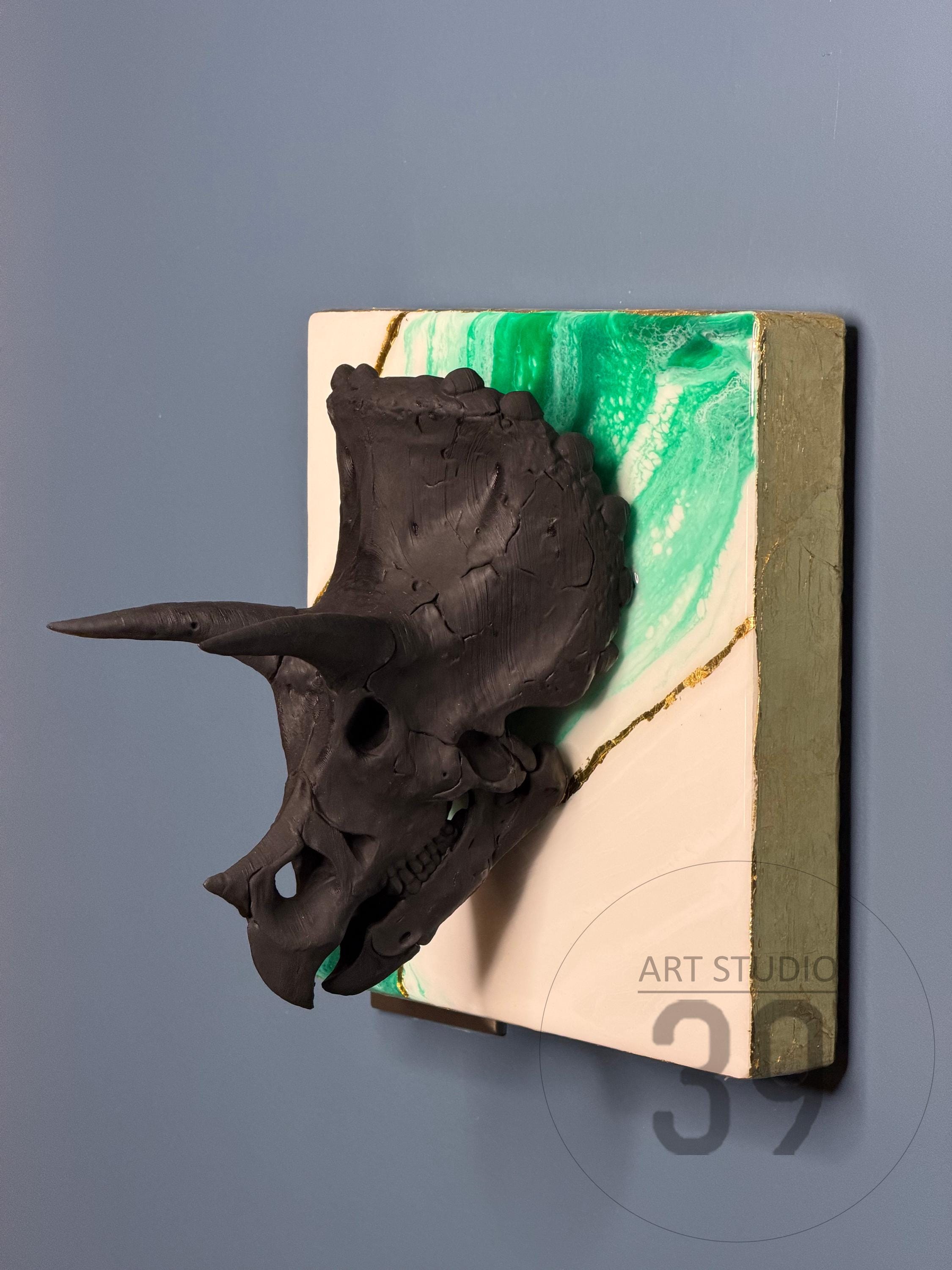 A black triceratops skull sculpture mounted on a square canvas. The canvas features a white and green marbled design with gold accents. The artwork is labeled "ART STUDIO 39".