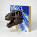 A dark brown dinosaur skull sculpture mounted on a square canvas. The canvas features a blue and white marbled design with gold accents. The sides of the canvas are painted gold. The sculpture is a unique piece of art.