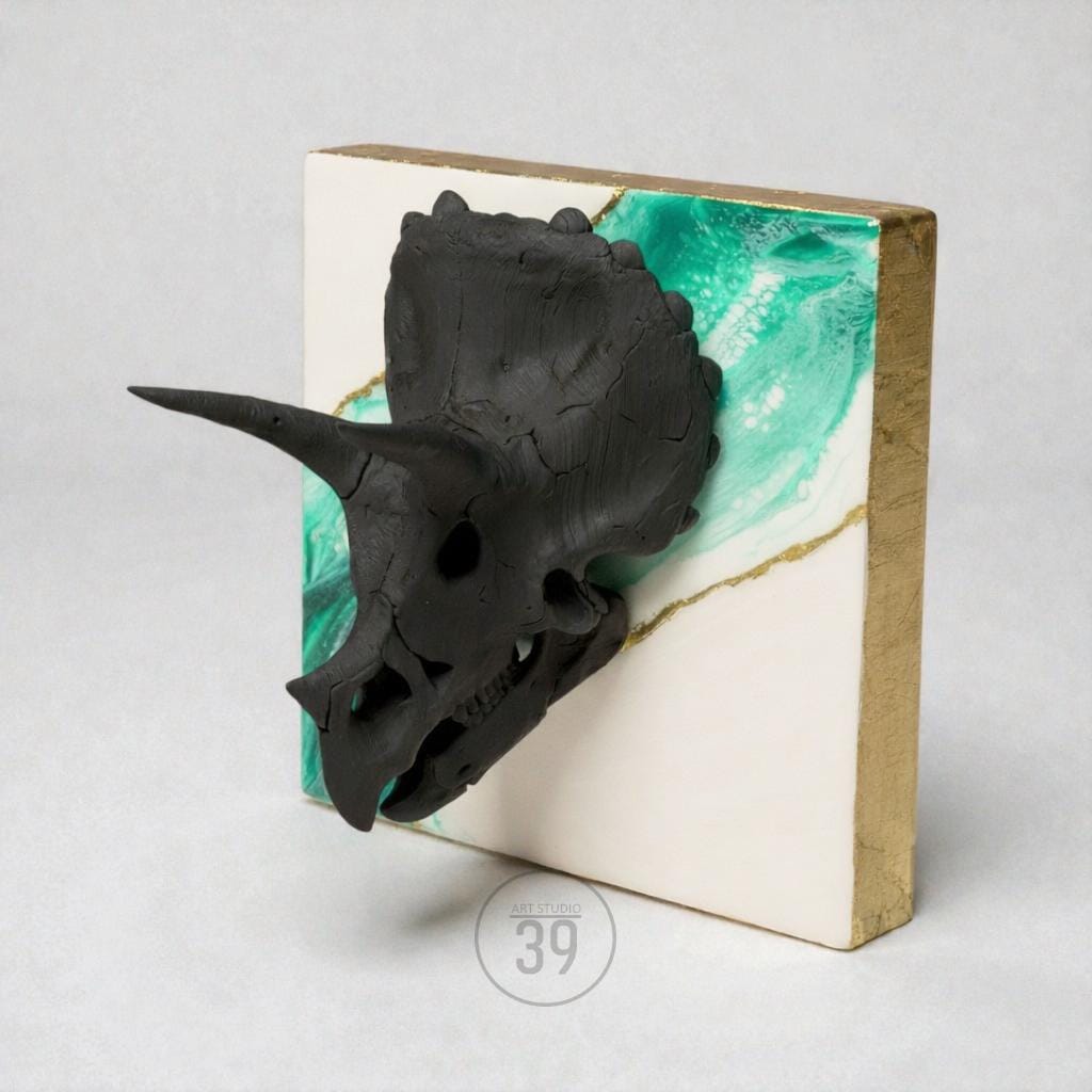 A black triceratops skull sculpture mounted on a square, white background with teal and gold accents. The edges of the square are gold. The sculpture is a unique piece of art.
