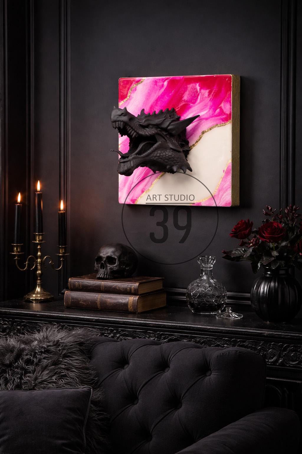 A black dragon sculpture mounted on a pink and white abstract art piece with gold accents. The artwork hangs above a black table with a skull, books, candles, and a decorative vase. The text "ART STUDIO 39" is visible.