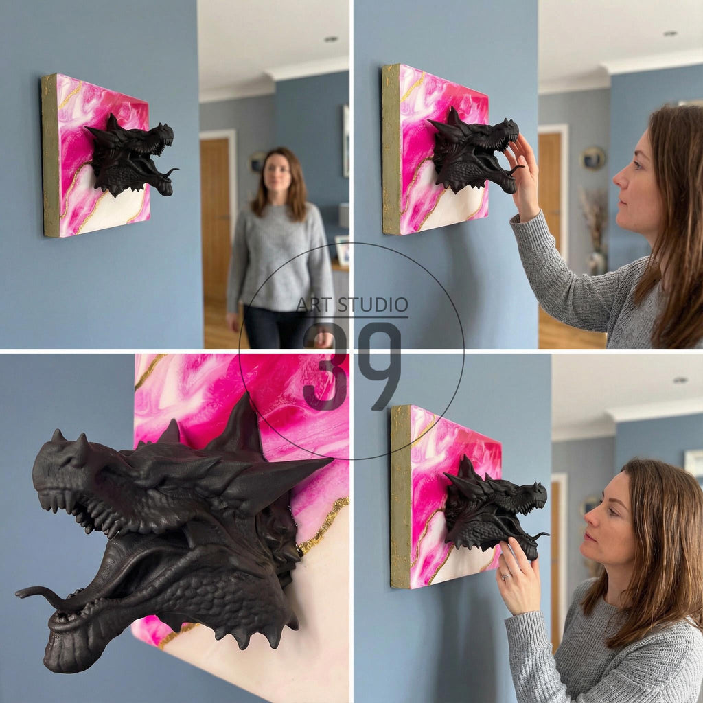 A black dragon head sculpture emerging from a square canvas with pink, white, and gold abstract art. The artwork is mounted on a light blue wall, showcasing a unique 3D effect. The dragon's open mouth and detailed scales are prominent.