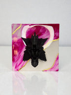 A black, detailed sculpture of a creature with wings, mounted on a square resin base. The base features a swirling design of pink, white, and gold accents. The sculpture is centered on the base.