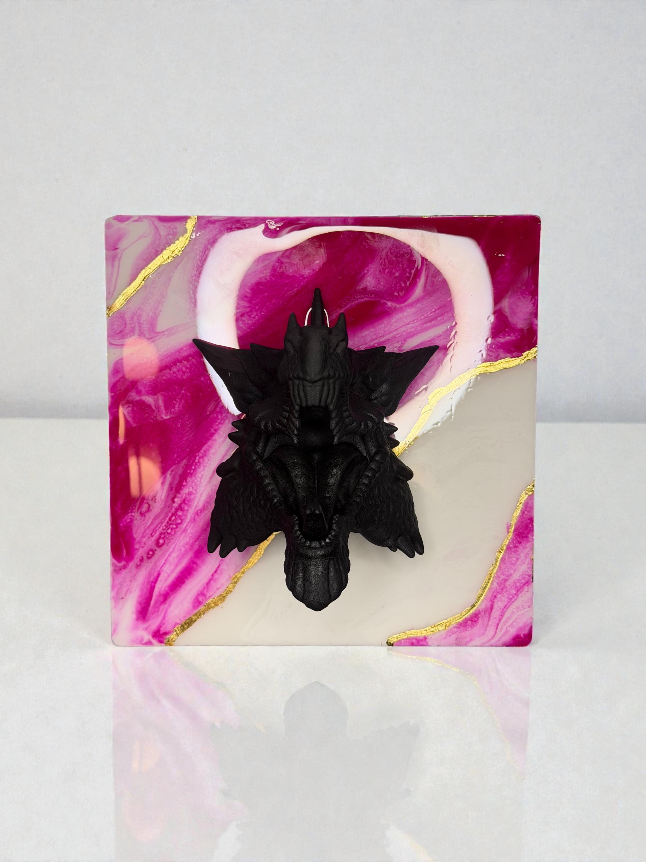 A black, detailed sculpture of a creature with wings, mounted on a square resin base. The base features a swirling design of pink, white, and gold accents. The sculpture is centered on the base.