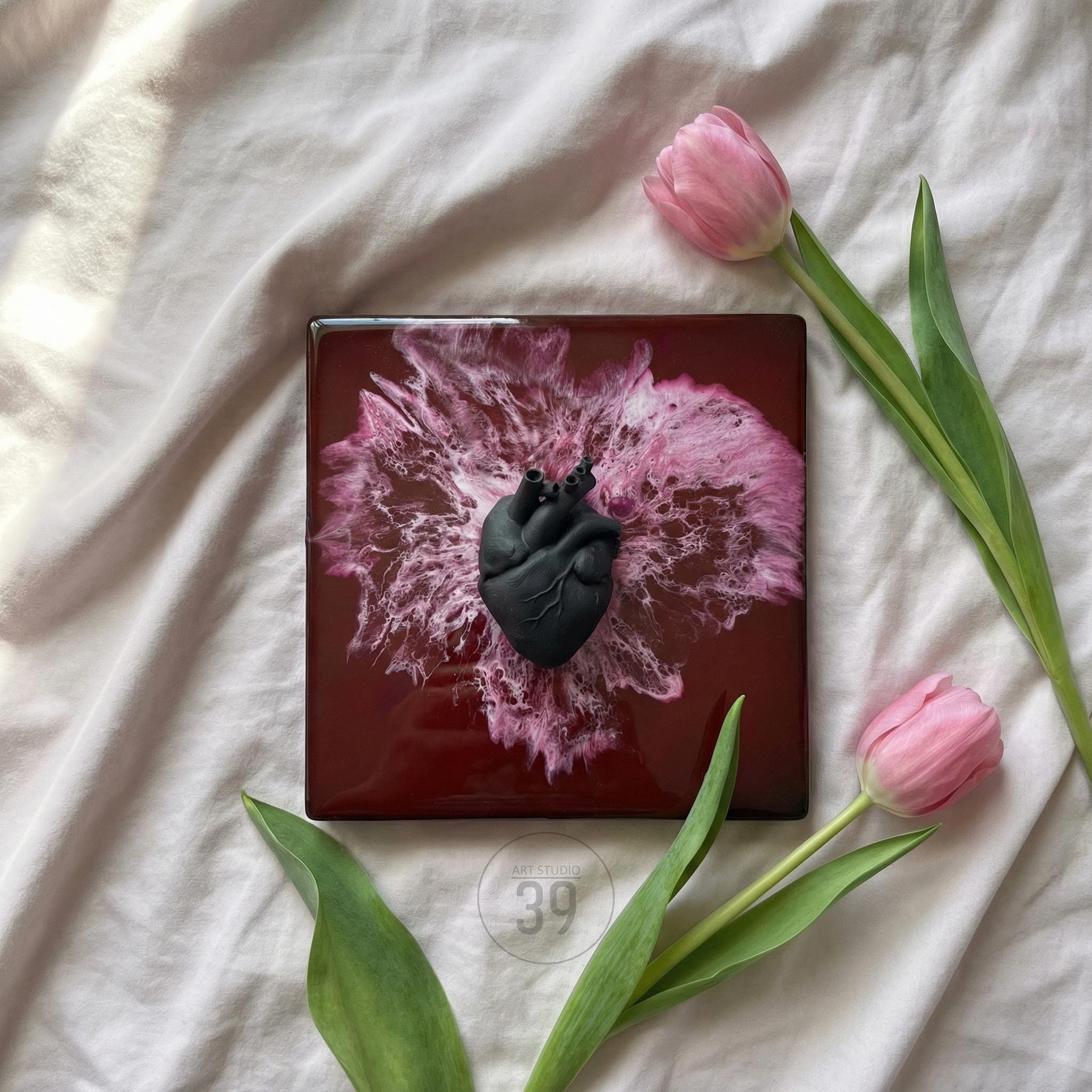 A square, dark red decorative tile featuring a black anatomical heart sculpture. The tile has a pink and white abstract design. Two pink tulips with green stems and leaves are arranged around the tile.