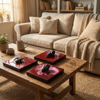 Three decorative square trays with dark red surfaces and wooden frames. Each tray features a different black sculpture: a spider, a bird skull, and a human heart. The trays are displayed on a rustic wooden coffee table.