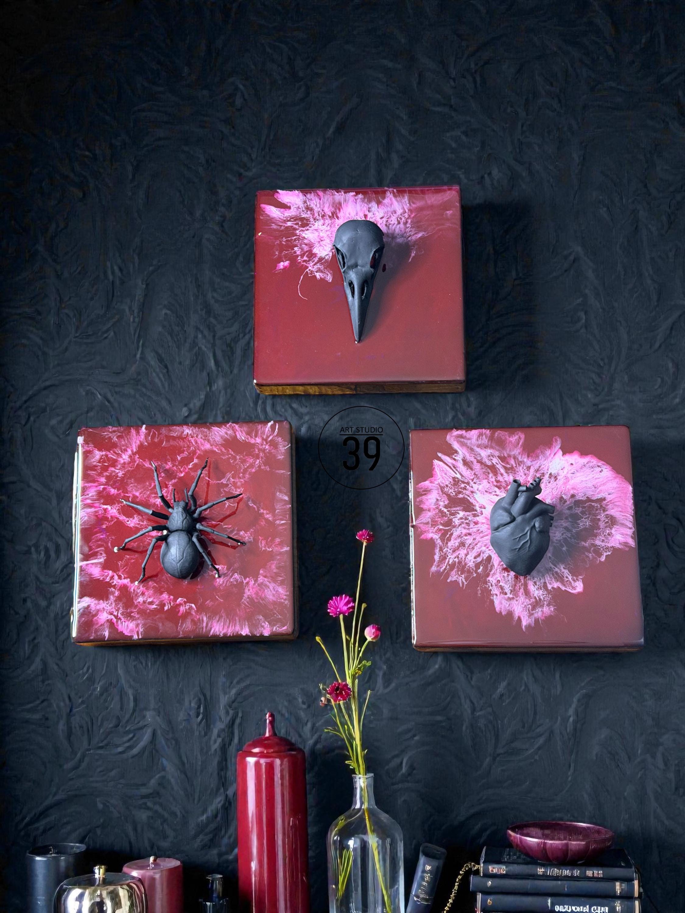Three square wall art pieces with dark red backgrounds and black sculptural elements. One features a bird skull, another a spider, and the third a heart. The art is displayed against a dark textured wall.