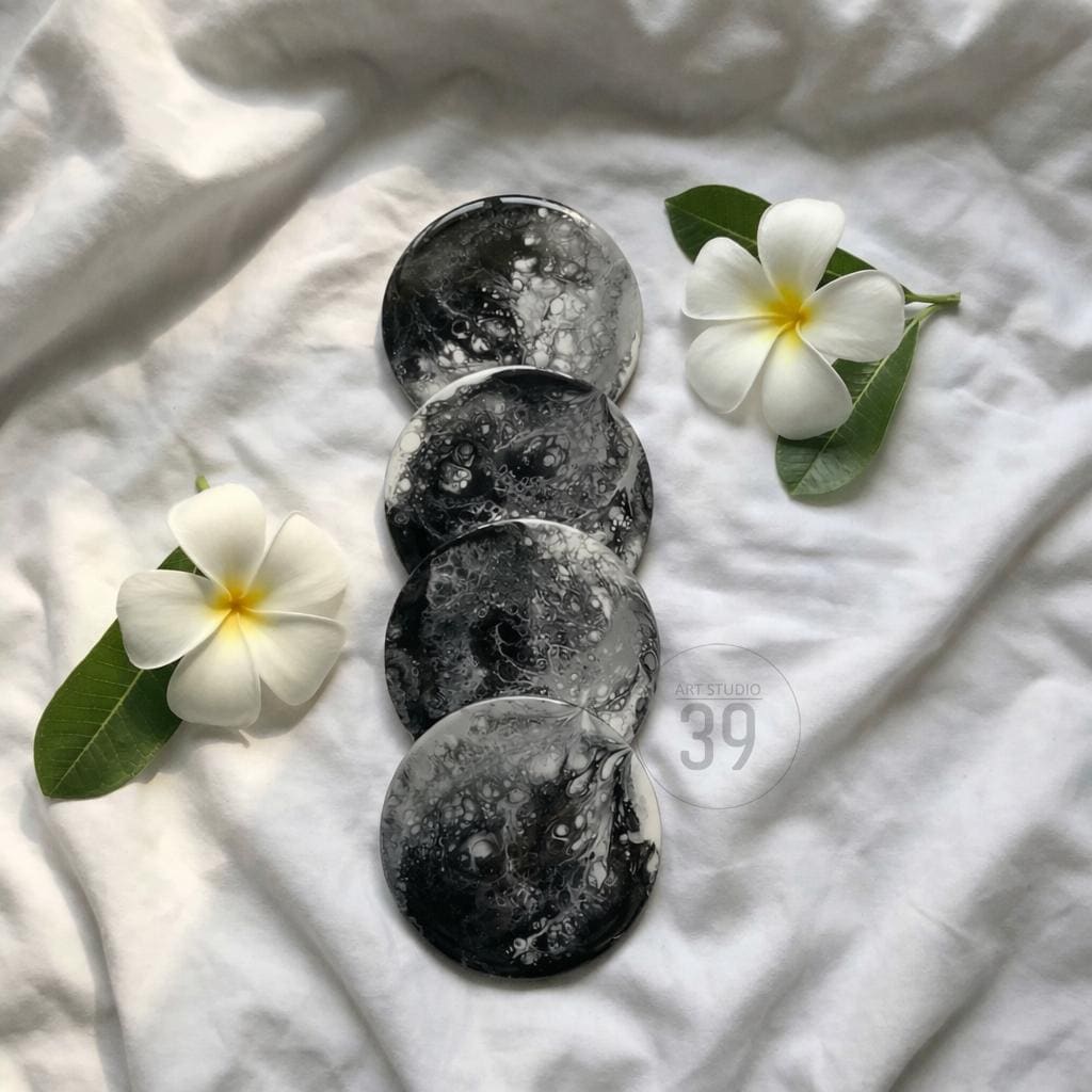 Four round coasters with a black and white marbled design are arranged on a white fabric surface. Two white plumeria flowers with yellow centers and green leaves are placed near the coasters. The coasters have a glossy finish.