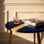 A navy blue round side table with gold accents and legs. A gold stripe crosses the table's surface. A glass of amber liquid and a book titled "Vogue: The Covers" rest on the table.