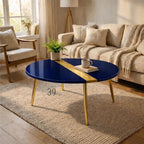 A navy blue oval coffee table with a gold stripe down the center. The table has gold legs and is set on a beige rug. A sofa, chair, and lamp are in the background. A mug and book are on the table.