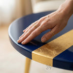 A close-up of a round, navy blue table with a gold leaf stripe. The table has gold-colored legs. Several gold rings adorn a hand resting on the table. The text "ART STUDIO 39" is visible.