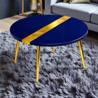 A round coffee table with a navy blue top and gold accents. The table has a gold stripe across the top and gold legs. The table is sitting on a gray shag rug.