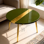 A round coffee table with a glossy olive green surface and a diagonal gold leaf accent. The table has gold-colored metal legs. The table is in a living room setting.