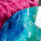 Close-up of a round, resin art piece with a swirling ocean-like design. The colors include shades of blue, turquoise, and white, resembling waves. The background is a soft pink, adding contrast to the artwork.