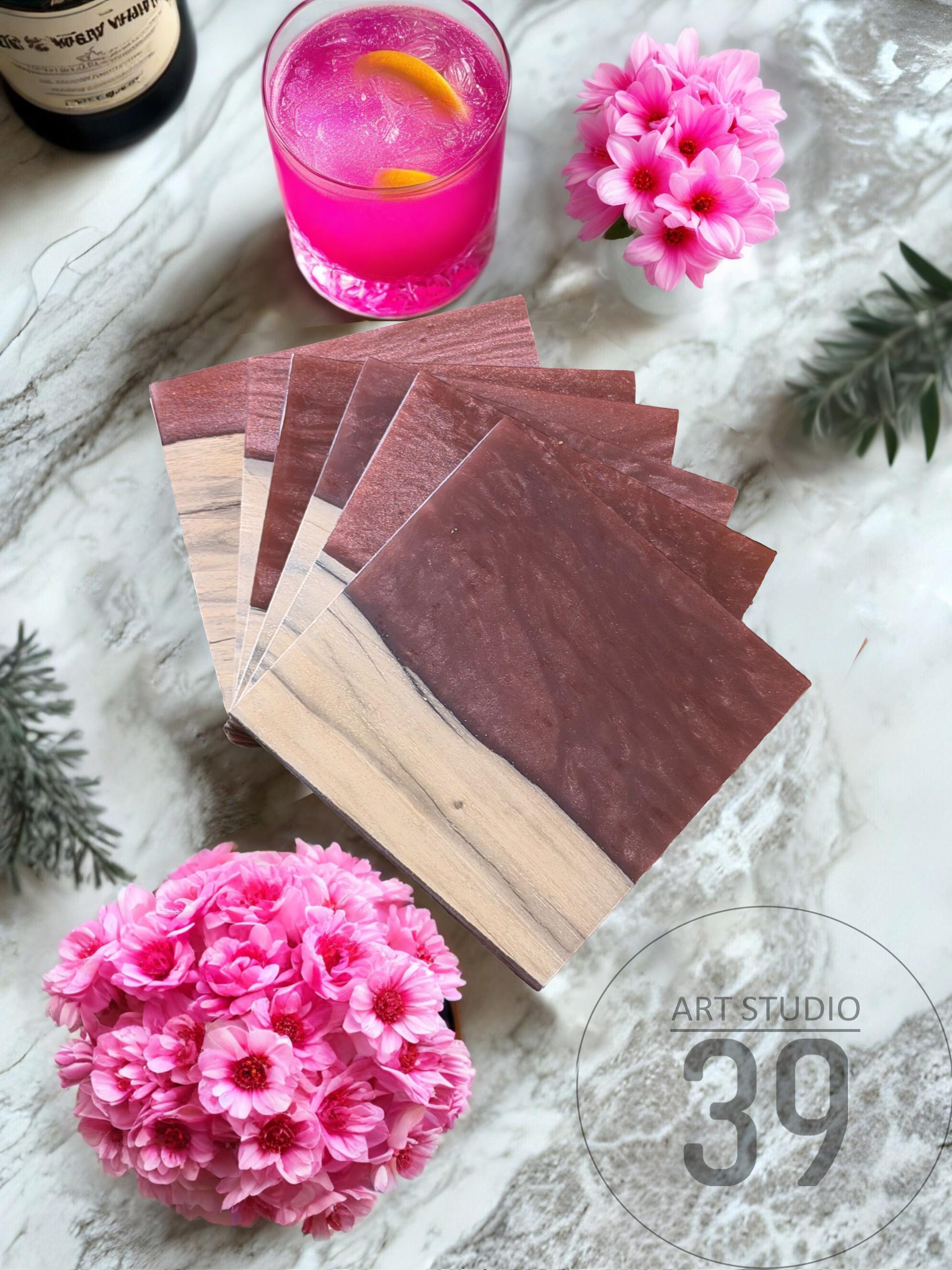 A stack of square wooden coasters with a two-tone design: light wood and dark brown. The coasters are arranged near a pink drink with a lemon slice, pink flowers, and the text "ART STUDIO 39".