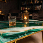 A green and white epoxy resin coffee table with gold accents. A lit candle lantern and a glass of amber liquid sit on the table. The table has gold legs and is in a dimly lit room.