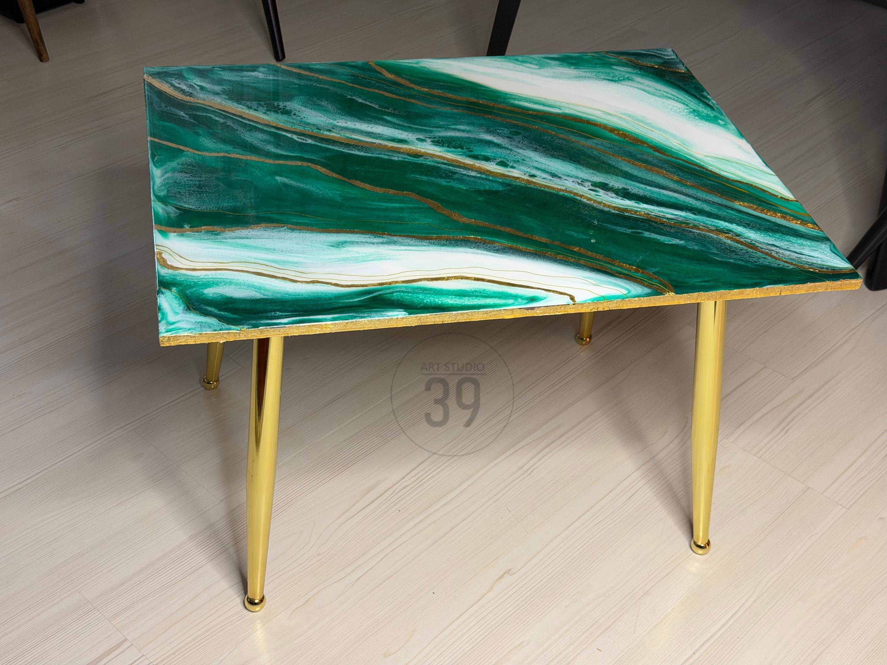 A rectangular coffee table with a green and white marbled design and gold accents. The table has four gold-colored, tapered legs. The top of the table has a glossy finish. The table is a modern home decor item.